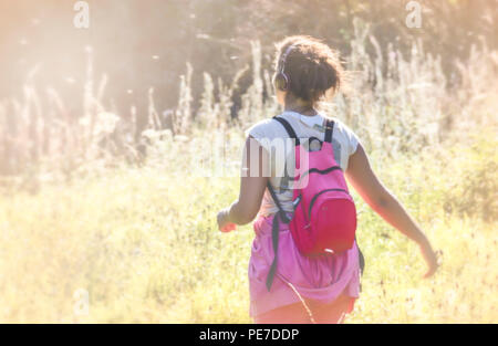 Eine junge Frau mit pinkem Rucksack und Kopfhörern, die während der Hitzewelle durch eine neblige Sommerwiese schlendert Stockfoto