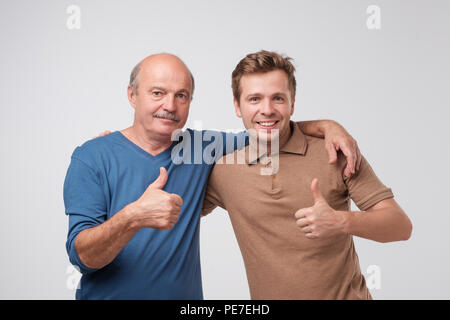 Porträt einer zwei glückliche junge Männer mit Daumen hoch über grauer Hintergrund isoliert. Reifen Sohn und Vater sind, mit einem guten, familiären Veranstaltung zufrieden Stockfoto