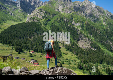 Junge Frau mit Rucksack steht auf einem Felsen mit erhobenen Händen und zu einem Tal unter suchen. Stockfoto