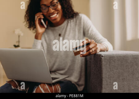 Frau in modischen zerrissenen Jeans auf dem Sofa zu Hause sitzen Gespräch am Handy mit einer Kreditkarte. Frau, die on-line-Zahlung auf Ihrem Laptop compu Stockfoto