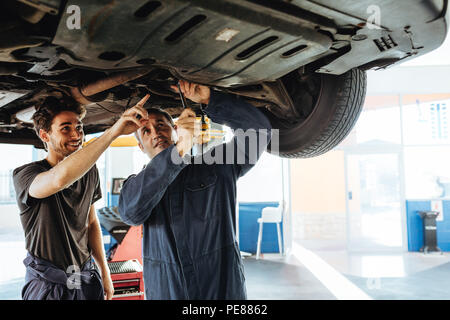 Mechaniker zur Festsetzung der Auto mit Kollegen zeigen und lächelnd. Zwei Auto Reparatur Männer unter einer angehobenen Fahrzeug in der Garage arbeiten. Stockfoto