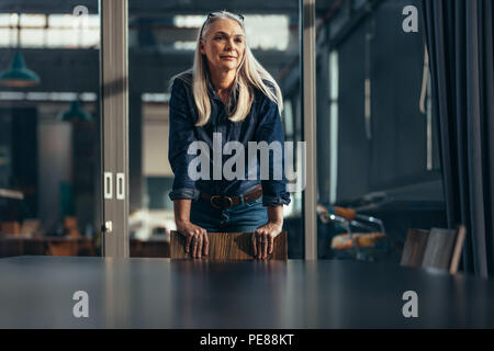 Portrait von erfolgreiche Geschäftsfrau gestützt auf einen Stuhl im Gemeinschaftsraum und Wegsehen. Ältere weibliche Unternehmer im Konferenzraum. Stockfoto