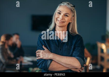 Portrait von zuversichtlich Senior Business im Amt stehende Frau mit verschränkten Armen. Reife Frauen im Amt mit Kolleginnen und Kollegen diskutieren, arbeiten an der b Stockfoto