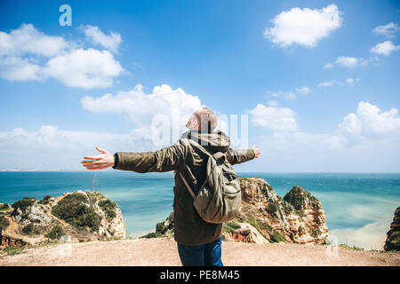 Ansicht von der Rückseite. Ein junger Mann tourist genießt die schöne Aussicht auf den Atlantischen Ozean und die Landschaft an der Küste in Portugal und hebt seine Hände nach oben zeigen, wie glücklich er ist. Stockfoto