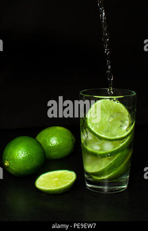 Kalte detox Wasser mit Eis und frischen Reifen Scheibe grün Limes auf schwarzem Hintergrund. Erfrischende Sommer und Getränke Konzept. Kalte detox Wasser mit Eis und Stockfoto
