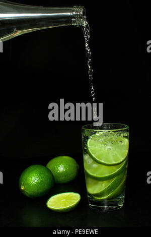 Erfrischende Sommer und Getränke Konzept. Strom der kaltes Wasser aus einer Flasche in ein Glas mit Eis und frischen Reifen Scheibe grün Limes auf Schwarz gegossen Stockfoto
