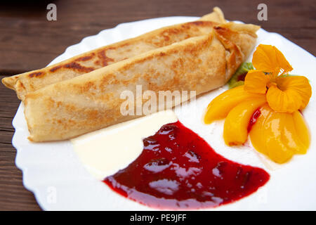 Weiße Platte mit Pfannkuchen Pfannkuchen mit Marmelade, Creme, Obst und Blumen auf einem Holztisch Stockfoto