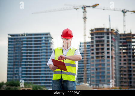 Architekten auf der Baustelle auf seine Armbanduhr mit Gebäuden und Krane im Hintergrund Stockfoto