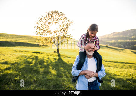Ein älterer Großvater eine kleine Enkelin eine piggyback Ride in der Natur. Stockfoto
