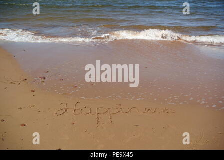 Verschwinden Wort "Glück" wunderschön handschriftlich auf dem roten Sand auf einem Strand mit einer Welle, die in der linken oberen Ecke, Prince Edward Island Stockfoto