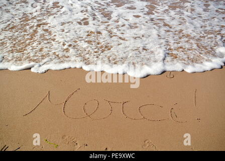 Eine schöne Dankbarkeit Nachricht 'merci' handschriftlich in einer kursiven Stil auf dem roten Sand auf einem Strand mit einer Welle, die hereinkommen, Prince Edward Island. Stockfoto