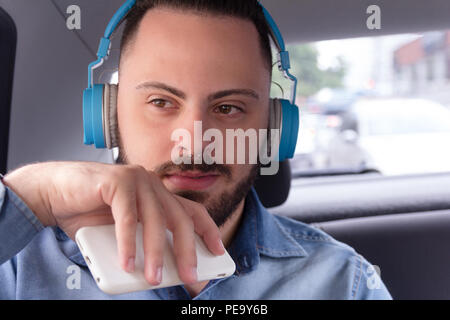 Glückliche junge bärtige Mann tragen Jeanshemd, auf ein Auto hören von Musik auf einem Smartphone. Konzept der Pendeln, Bestrebungen, das Leben in der Stadt, Konzentration, Jo Stockfoto