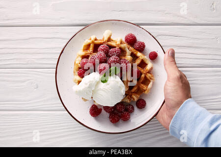 7/8 Schuß von Mann Halteplatte der belgische Waffeln mit Himbeeren und Eis über weiße Holztisch Stockfoto