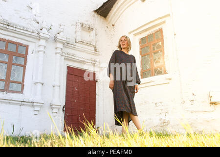 Junge schöne Modell Frau in langen vintage Kleid im Stil Boho mit Blumenmuster steht auf der grünen Wiese in der Nähe von weißen Stein alte Wand mit Fenstern und Stockfoto