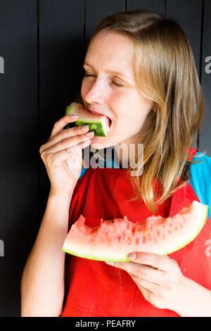 Eine schöne und junge Mädchen beißt eine frische, leckere und saftige Wassermelone. Sie ist sehr hungrig und will es zu essen. Wassermelone, sie brach in zwei kleinen Stockfoto