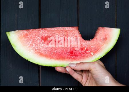 Ein Stück saftige, frische Wassermelone ist in einer Hand auf einem hölzernen Hintergrund, die in schwarzem Lack lackiert gehalten wird. Stockfoto