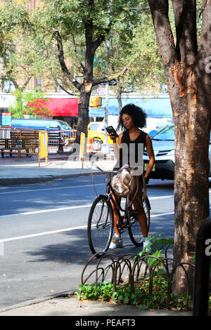 NEW YORK, NY - 31. August: junge schwarze Frau in einem eleganten Kleid überprüft Ihr Telefon beim Sitzen auf dem Fahrrad auf Bleecker Street, Manhattan am 31. August Stockfoto