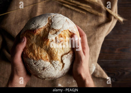 7/8 shot der männlichen Baker holding Brot über Holztisch mit Weizen und Sack Stockfoto