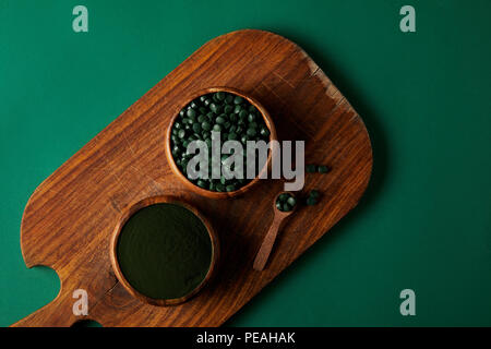 Blick von oben auf ein Schneidbrett, Löffel aus Holz, Schalen mit Spirulina Pulver, spirulina in Pillen am grünen Tisch Stockfoto