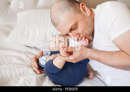 Berühren Portrait von mittleren Alter kaukasischen Vater im weißen T-Shirt im Bett liegend mit neugeborenen Sohn küßte seine Füße Zehen, parenting Kindheit bondin Stockfoto