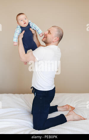 Portrait von mittleren Alter kaukasischen Vater im weißen T-Shirt und schwarze Jeans sitzen auf dem Bett zuhause, Holding, die neugeborenen Sohn macht ihn Lachen, Stockfoto