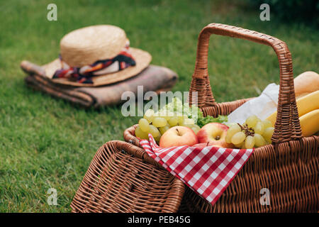 Köstliche Früchte im Korb auf grünem Gras im Picnic, Hut und Decke auf Hintergrund Stockfoto