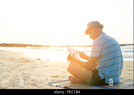 Älterer Mann bei der Arbeit auf seinem Laptop am Strand bei Sonnenuntergang, Freiberufler Konzept in Reisen Stockfoto