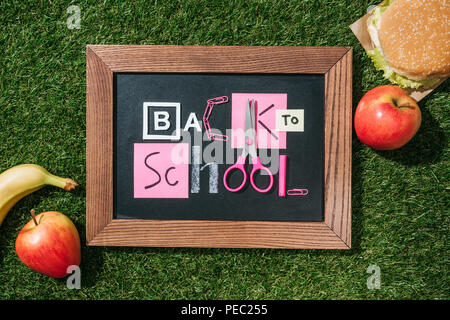 Flach mit frischen Äpfeln, Bananen, Burger und Tafel mit zurück in die Schule Schriftzug aus rosa und weiße Objekte auf grünen Rasen Stockfoto