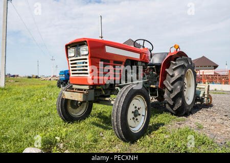 Einen kleinen roten Traktor steht auf einem Hof auf grünem Gras und wartet auf Arbeit zu beginnen. Stockfoto