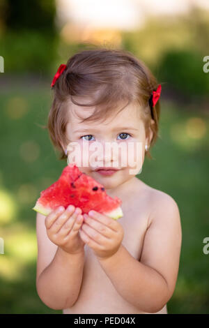 Closeup Portrait von niedlichen kleinen Mädchen, Essen, Wassermelone auf dem Gras im Sommer gesunde Snack für Kinder. Kleines Mädchen im Garten spielen holdin Stockfoto