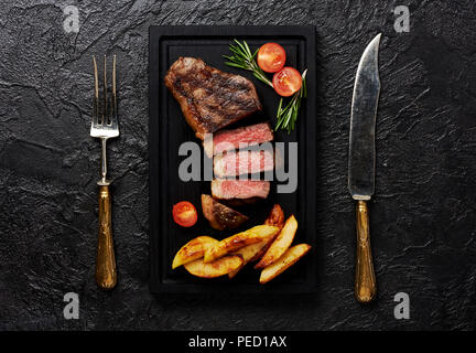 Fleisch Picanha Steak, traditionelle brasilianische Schnitt mit Tomaten, Kartoffelecken und Rosmarin auf schwarzes Fleisch Schneidebrett. Steak mit Messer und Gabel. Schwarz konkreten Hintergrund. Stockfoto