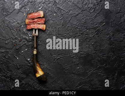 Scheiben von medium gegrilltes Steak auf Fleisch Gabel mit Salz und Pfeffer auf Schwarz konkreten Hintergrund. Kopieren Sie Platz. Ansicht von oben. Stockfoto