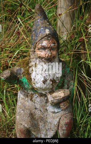 Sehr alt und Wetter getragen Gartenzwerg mit weißem Bart, Moos bedeckt Jacke und Schlamm Abdeckung hat Holding und Amboss und Hammer hingewiesen Stockfoto