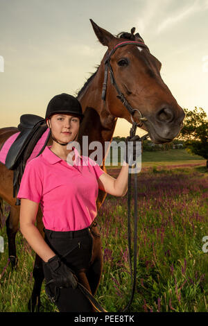 Schöne lächelnde Mädchen Jockey stand neben ihr braunes Pferd Tragen besonderer Uniform auf einen Himmel und grünen Feld Hintergrund auf einen Sonnenuntergang. Stockfoto