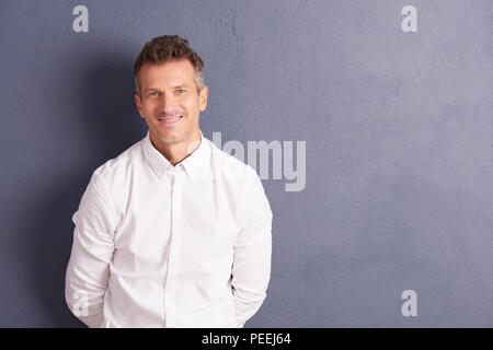 Stattlich im mittleren Alter Mann mit Hemd und Suchen an der Kamera, während an der grauen Wand stehend. Stockfoto