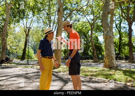 Bunt gekleidete italienische Männer, beide mit Strohhüten, treffen sich und begrüßen sich an einem Sommermorgen in einem baumbestandigen Park. Piacenza, Italien. Stockfoto