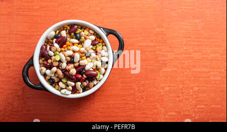 Gesunde Ernährung Konzept. Sortiment von Hülsenfrüchten in Emaille Schüssel auf orangem Hintergrund, isoliert, Ansicht von oben, kopieren. Stockfoto