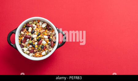 Gesunde Ernährung Konzept. Sortiment von Hülsenfrüchten in Emaille Schüssel auf rotem Hintergrund, isoliert, Ansicht von oben, kopieren. Stockfoto