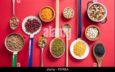 Gesunde Ernährung Konzept. nach oben Blick auf fflat lag der Sortiment von Leguminosen auf rot Tischplatte Hintergrund, in der Schaufel, Schüssel und Pfannen. Stockfoto