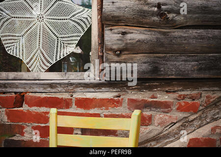 Detailansicht über einen Teil eines alten Holzhaus, Fenster mit Stickerei auf der einen Seite, alte verwitterte Holzbohlen auf anderen und Backsteine in der Unterseite. Pa Stockfoto