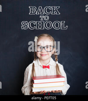 Süße kleine Mädchen Schüler holding Stapel Bücher und lächelnd. Schülerin in Gläsern auf blackboard Hintergrund. Zurück zu Schule. Stockfoto