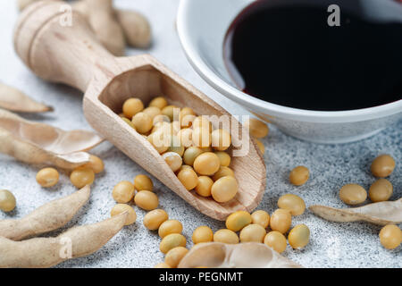 Sojabohnen in Löffel aus Holz, Hülsen und Sojasauce in einer weißen Schüssel auf einem grauen Betonflächen. Selektiver Fokus Stockfoto