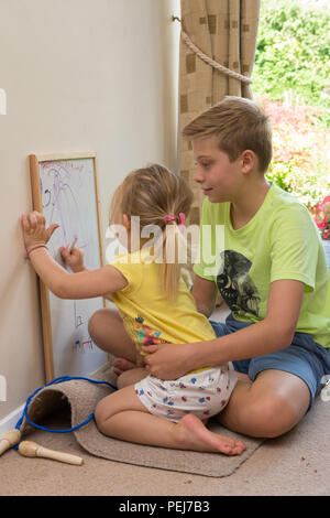 Junge zwei Jahre altes Mädchen sitzt auf dem Schoß der jungen 12-jährigen Bruder, Zeichnung mit Farbstift auf dem Whiteboard, künstlerische, Stockfoto