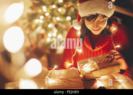 Frohe Weihnachten. Mädchen Verpackung Weihnachtsgeschenke in Lichter am Abend festliches Zimmer unter Baum Beleuchtung. Mädchen in santa hut öffnen Moderne Handwerk gi Stockfoto