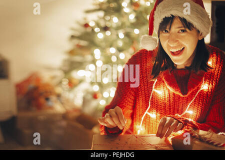 Happy girl in Santa Hat und Red Pullover Verpackung Weihnachtsgeschenke in Lichter am Abend festliches Zimmer im Baum Beleuchtung. Öffnen moderne craft Geschenke Stockfoto