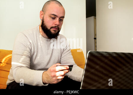 Geschäftsmann von zu Hause aus zu arbeiten und die Zahlung der Rechnungen per Kreditkarte über Laptop. Mann mit einem Laptop beim Sitzen auf einem Sofa in seinem Wohnzimmer. Stockfoto