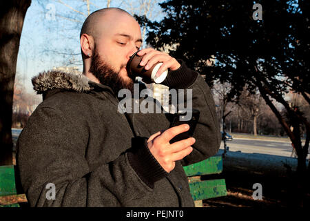Junger Mann im City Park sitzen mit Smart Phone und trinken Kaffee zum Mitnehmen. Hübscher Kerl im Herbst Outfit entspannend. Stockfoto