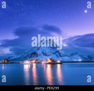 Fischerboote in der Nähe von Pier am Meer gegen Snowy Mountains und starry lila Himmel mit Mond in der Nacht auf den Lofoten Inseln, Norwegen. Winterlandschaft mit Stockfoto