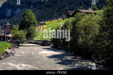 Malerische alpine Szene mit einem Fluss, einige typische Chalets aus Holz und eine gelbe Zug, den Berg hinunter. Grindelwald, Berner Oberland, Schweiz Stockfoto