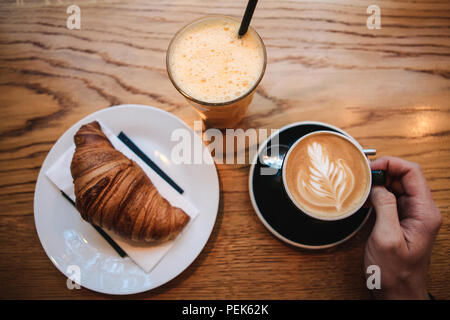 Ansicht von oben. Ein Mann nimmt eine heiße Tasse duftenden Cappuccino. In der Nähe des Tisches ist ein Croissant und ein Glas mit frischem Orangensaft. Frühstück am Morgen. Stockfoto